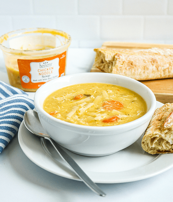 A Bowl of Heinen's Chicken Noodle Soup Beside an Open Plastic Soup Container and a Baugette