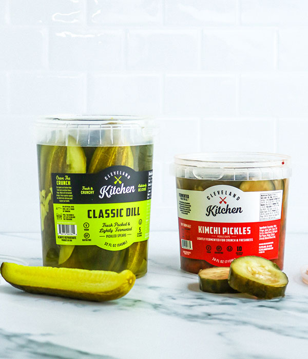 Two Open Containers of Cleveland Kitchen Pickles with a Whole Pickle Spear and Two Pickle Chips Sitting Beside the Containers
