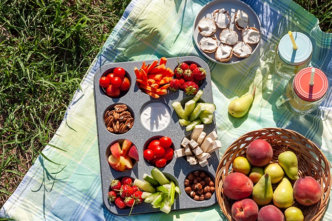 Summertime healthy picnic with fresh vegetables, nuts and fruits served in the muffin pan; refreshing drink in mason jar and some snack on blue cloth in the garden.