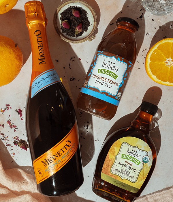 A Bottle of Non Alcoholic Sparkling Wine, A Bottle of Heinen's Unsweetened Iced Tea and a Bottle of Heinen's Pure Organic Maple Syrup Laying Flat on a Neutral Surface with Fresh Fruit
