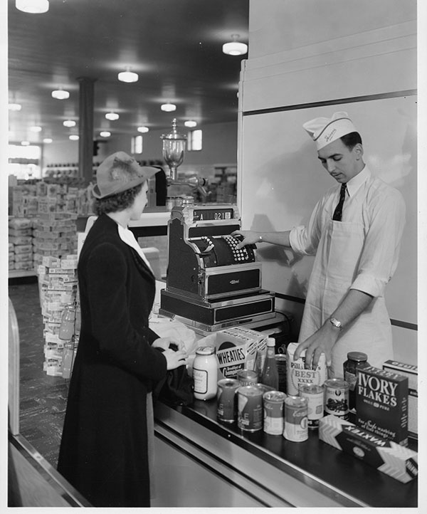 A vintage image of a woman purchasing groceries from a Heinen's grocery store with the help of the employee running the cash register.