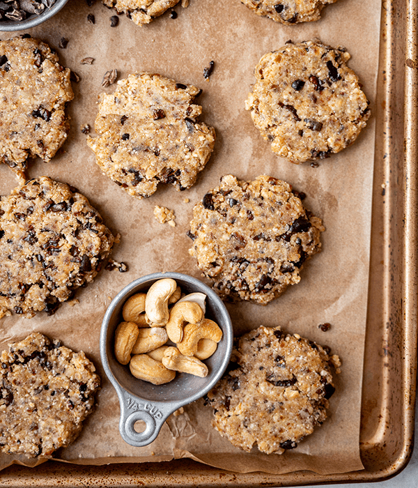 Several No Bake Chocolate Chip Cookies on a Parchment Lined Baking Sheet with a Cup of Cashews