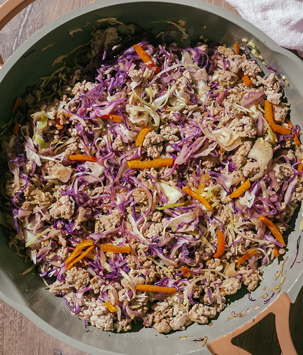 Overhead of pork, cabbage, and coleslaw mix being fried in a pan