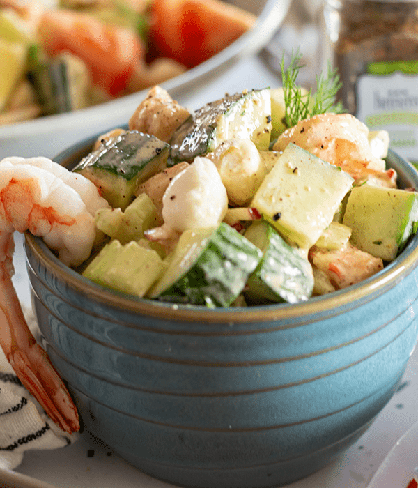 Close Up Shot of a Cold Citrus Shrimp Salad in a Small Blue Bowl with a Full Piece of Shrimp Hanging Over the Side