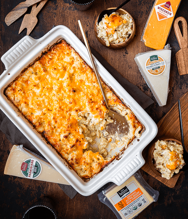 Five Cheese Baked Macaroni and Cheese in a White Baking Dish with a Metal Serving Spoon Surrounded by Packages of Heinen's Hand-Selected Cheeses