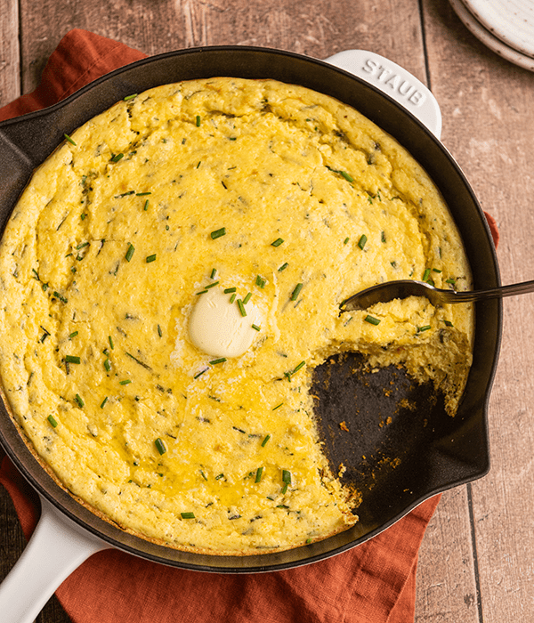 Cheddar Chive Spoon Bread in a Cast Iron Skillet with a Serving Spoon