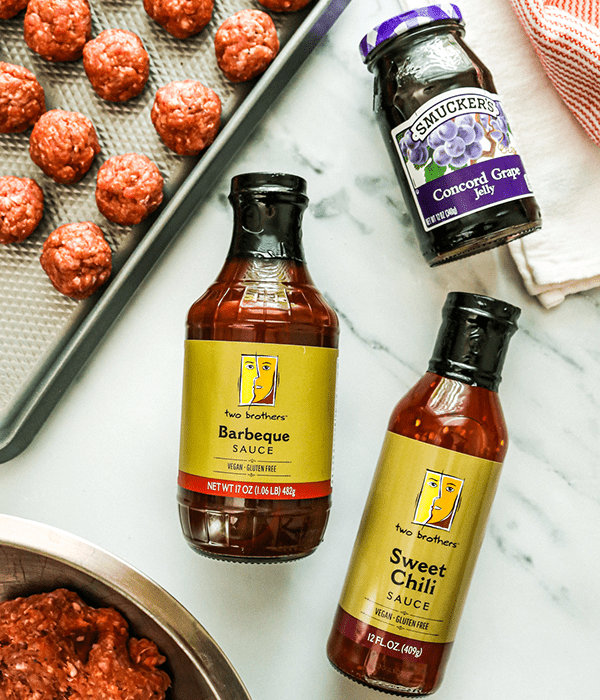 Bottles of Two Brothers Barbecue Sauce, Two Brothers Sweet Chili Sauce, and Smucker's Concord Grape Jelly on a Counter Beside a Tray of Uncooked Meatballs