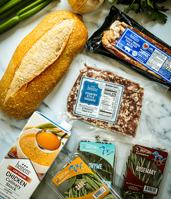 An Assortment of Fresh Ingredients including Sourdough Bread, Bacon, Ground Sausage, Chicken Stock, and Fresh Herbs on a Marble Surface