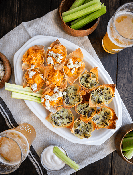 Daily Bites Spinach and Artichoke and Buffalo Chicken dips in baked wontons served on a plate with celery and blue cheese crumbles. 