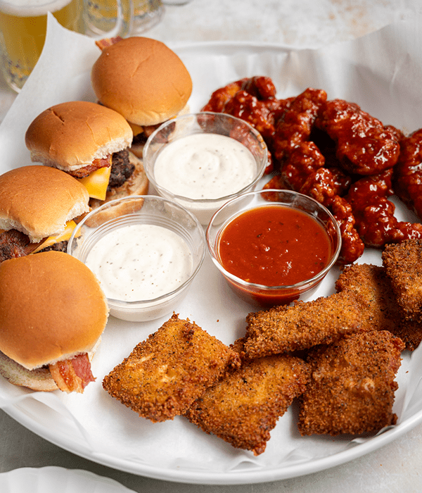 An Angled Image of a Copycat Triple Dipper Appetizer Platter Featuring Chicken Fingers, Cheeseburger Sliders, and Fried Mozzarella with Dipping Sauces