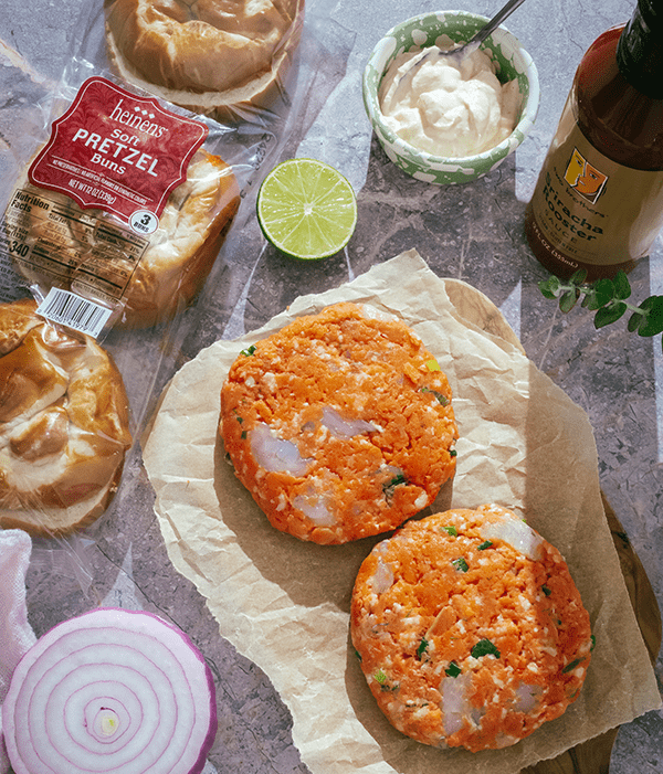 Heinen's Wild Salmon and Shrimp Burgers on parchment paper next to red onion, pretzel buns, and siracha rooster sauce. 