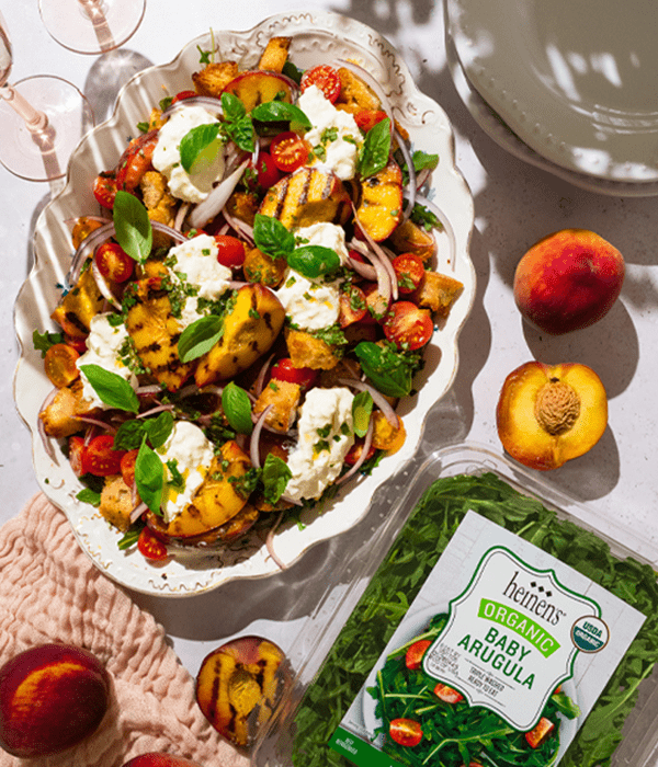 Plate with tomato, peach, and stracciatella salad, and a container of Heinen's Baby Arugula.